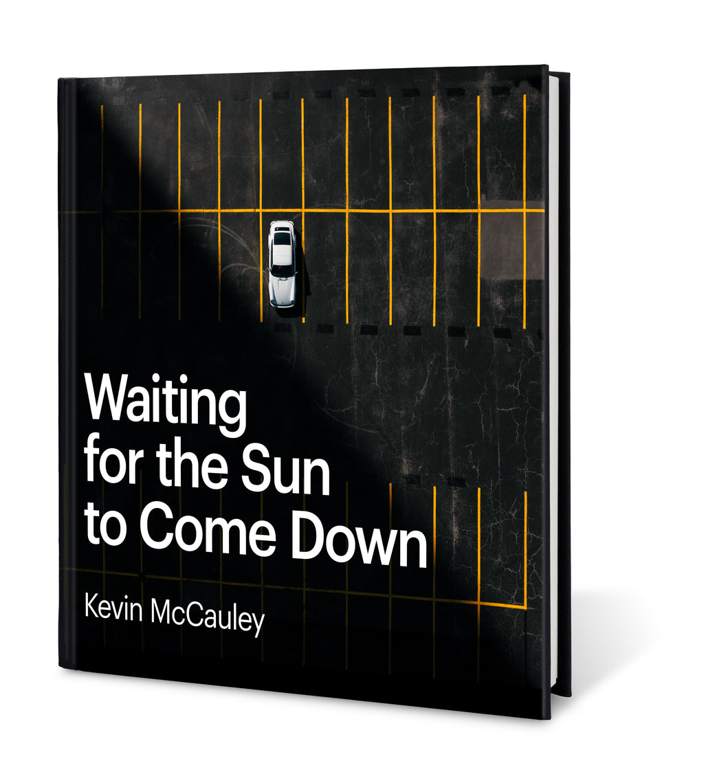 A hardcover book titled 'Waiting for the Sun to Come Down' by Kevin McCauley, shown at a slight angle on a white background. The cover is an overhead view of a white Porsche 911 with a long black shadow cut shockingly across the cover. Published by Carrara Media.