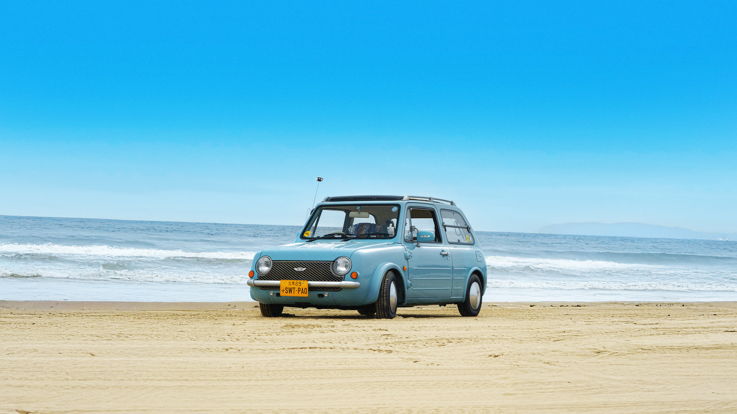 A classic blue Nissan Pao parked on the beach in Southern California, in an image from Bayside Magazine Issue 01, a new periodical celebrating coastal car culture. Published by Carrara Media.