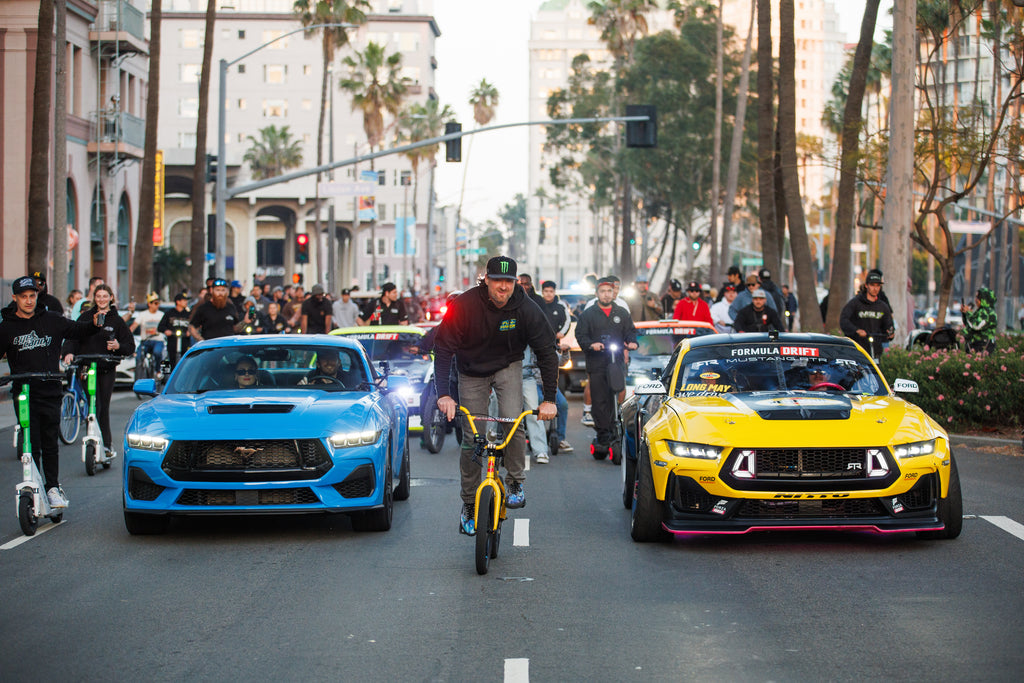 Image of Formula Drift cars on the street, with Vaughn Gittin Jr. of RTR Vehicles riding a bicycle between James Deane in Ford Mustang and Ben Hobson in Ford Mustang RTR FD, photo by Larry Chen. From the 400-page coffee table book Life At Shutter Speed by Larry Chen, foreword by Jeff Zwart and afterword by Magnus Walker. Published by Carrara Media.