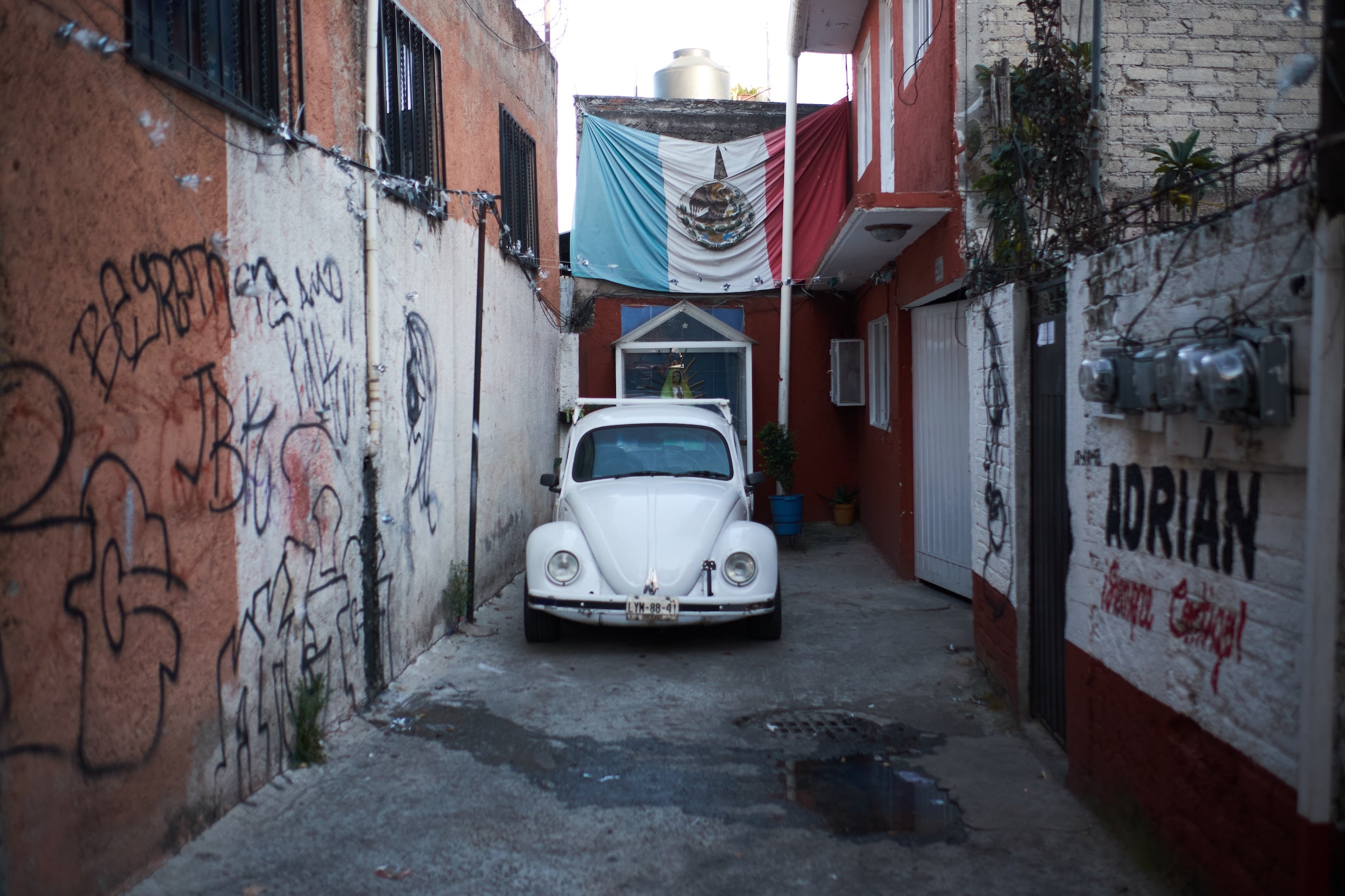 Image of a white Volkswagen Beetle parked in a red alleyway with a Mexican flag overhead, from the new book Vochos Unidos by Rodrigo Gaya Villar, published by Carrara Media. A coffee table book of automotive photography showing the Volkswagen Beetle across Mexico, aka "El Vocho." Limited Edition with slipcase and Leen Customs pin available for 500 copies only.