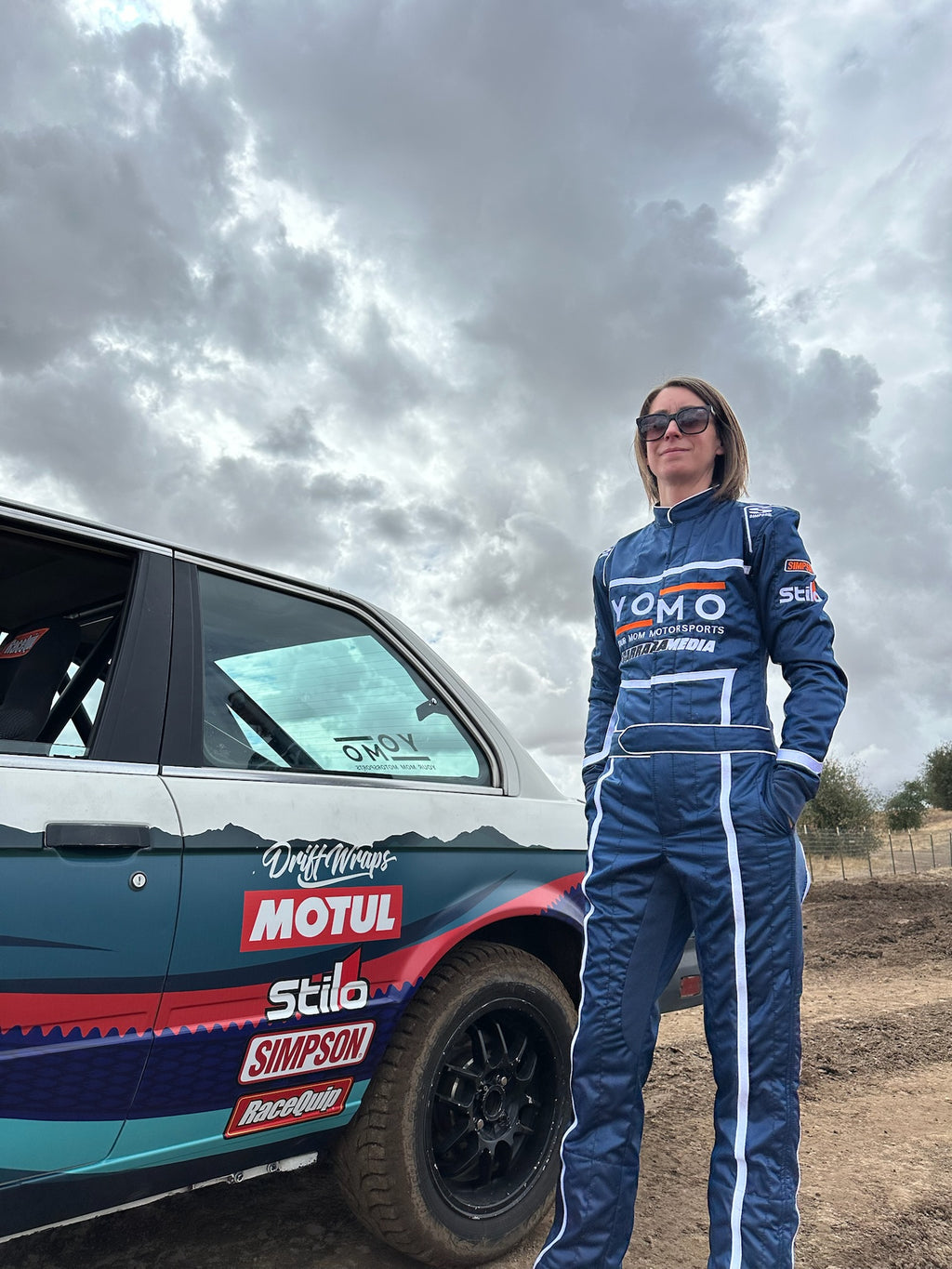 Image of Your Mom Races Rally author Kelly Conroy in her race suit with her BMW E30 rally car named “Nessie.” From the book Your Mom Races Rally, A nonfiction rally racing memoir describing Conroy’s inspiring journey to become a racecar driver to confront her depression after motherhood. Published by Carrara Media.