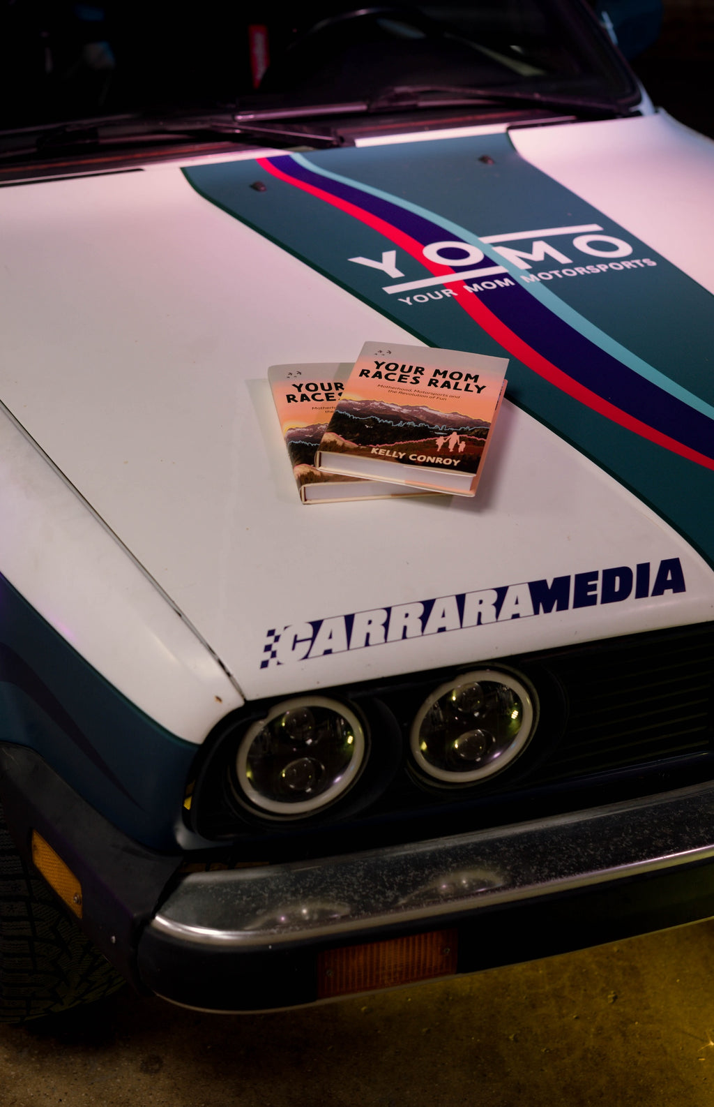 Image of the book Your Mom Races Rally by Kelly Conroy on the hood of her BMW E30 rally car named “Nessie.” A nonfiction memoir describing Conroy’s inspiring journey to become a racecar driver to confront her depression after motherhood. Published by Carrara Media.