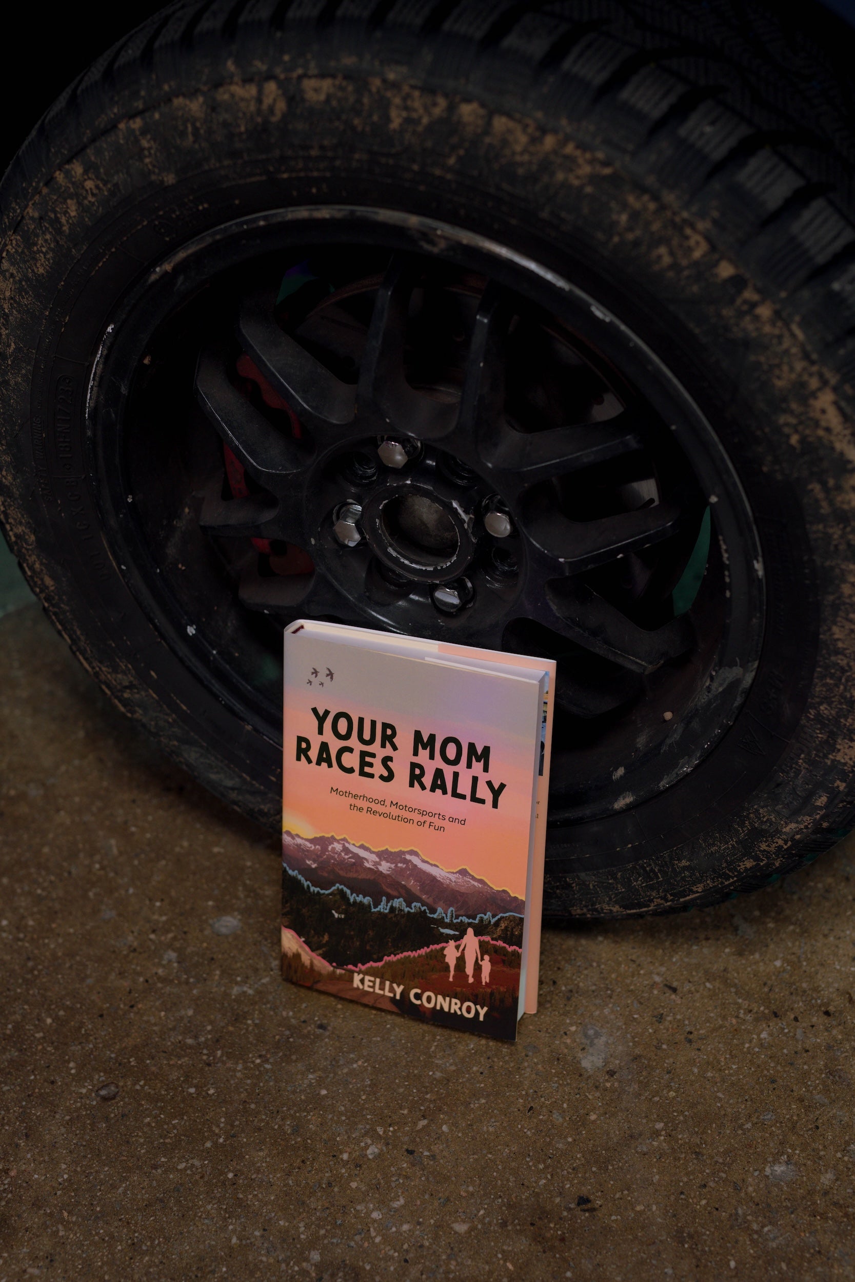 Image of the book Your Mom Races Rally by Kelly Conroy leaning against the wheel of her BMW E30 rally car named “Nessie.” From the book Your Mom Races Rally, a nonfiction rally racing memoir describing Conroy’s inspiring journey to become a racecar driver to confront her depression after motherhood. Published by Carrara Media.